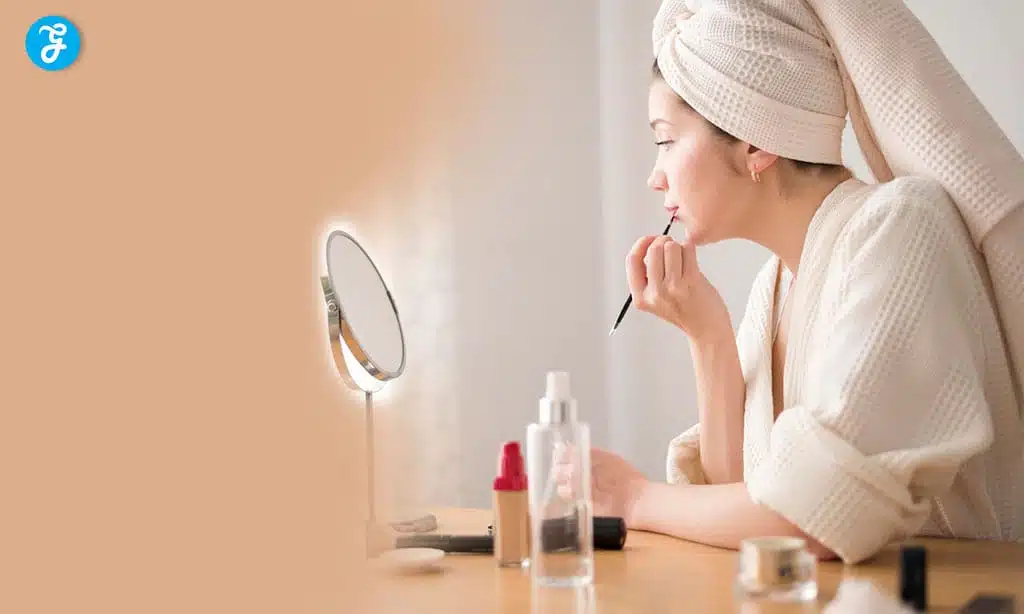 Woman applying makeup with towel on head in cozy bathroom.
