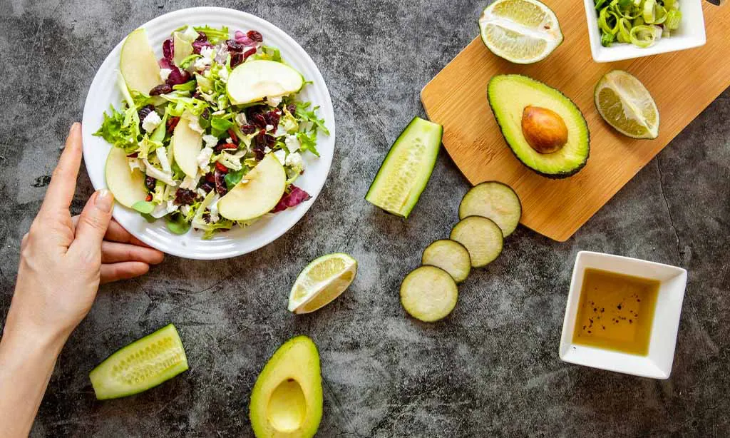 Fresh salad on a plate, surrounded by avocado, cucumber, lime, and a small bowl of dressing. A hand holds the plate