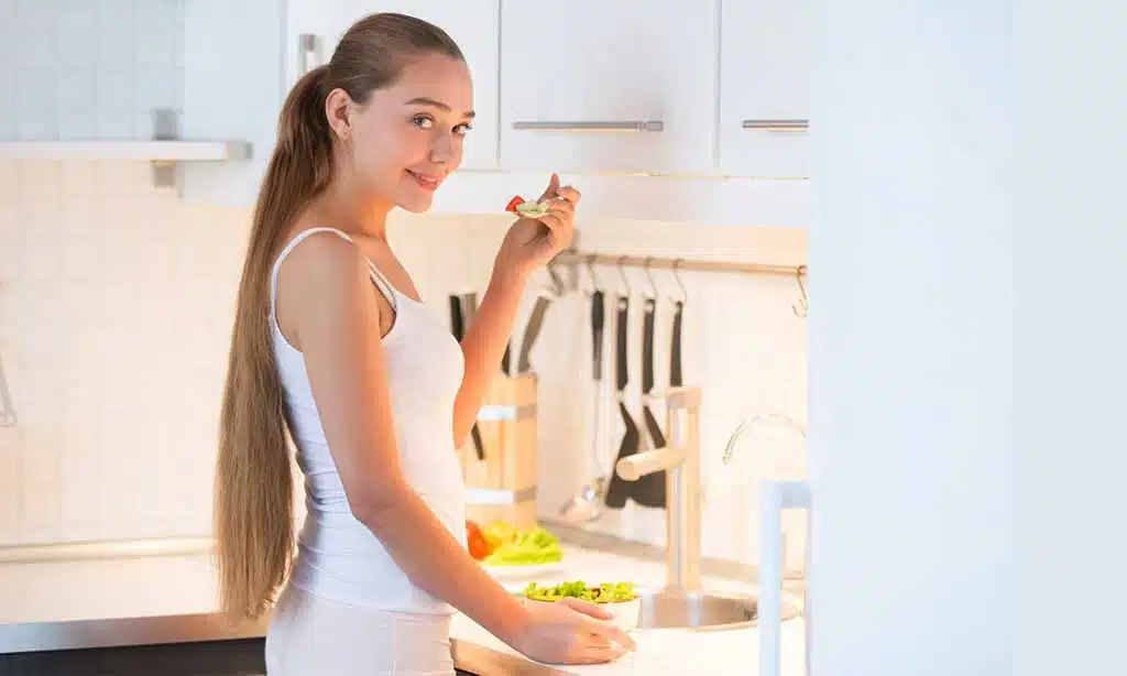 Young woman in a white outfit, smiling while holding a spoon and a bowl of salad in a modern, white kitchen