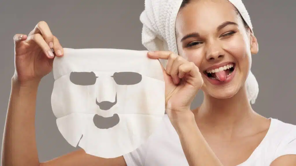 Woman with a towel on her head holding a face mask and sticking her tongue out