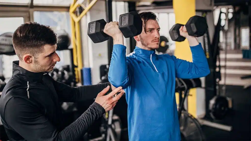 Trainer helping man with dumbbell shoulder press at gym.