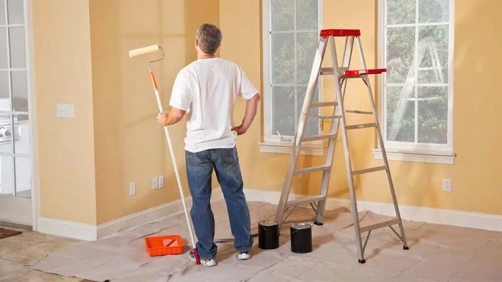 A man paints a room with yellow walls, ladder, and supplies ready