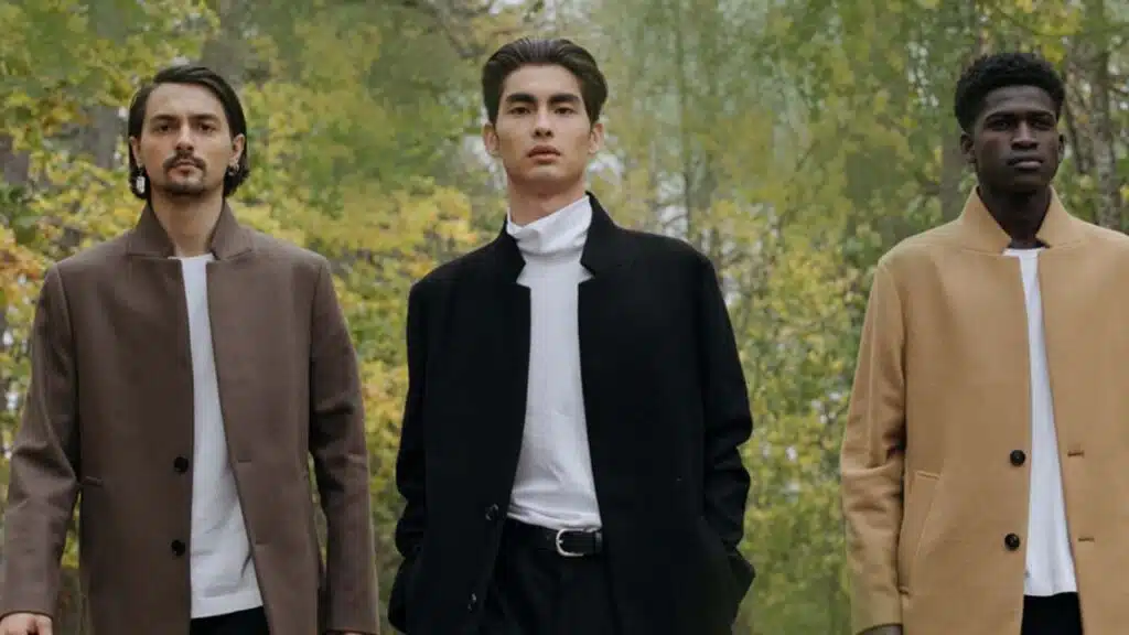 Three men in stylish coats walking outdoors in a wooded area.