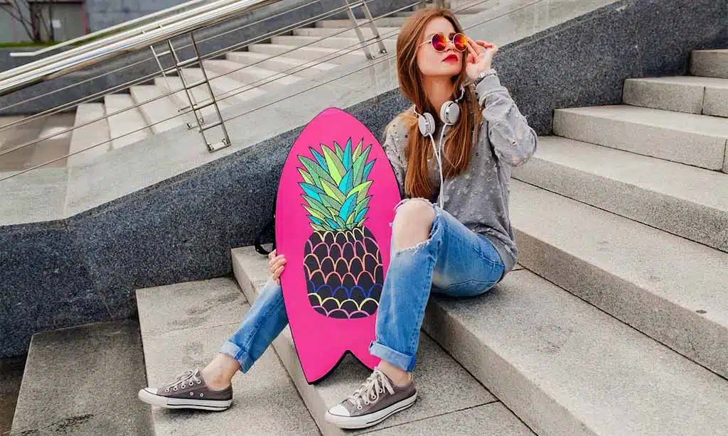A young woman sits on concrete steps, holding a pink skateboard with a pineapple graphic.