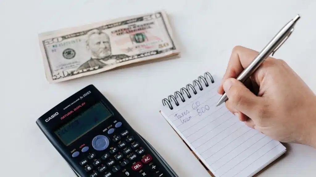 Person budgeting with a calculator, notepad, and cash on a white surface