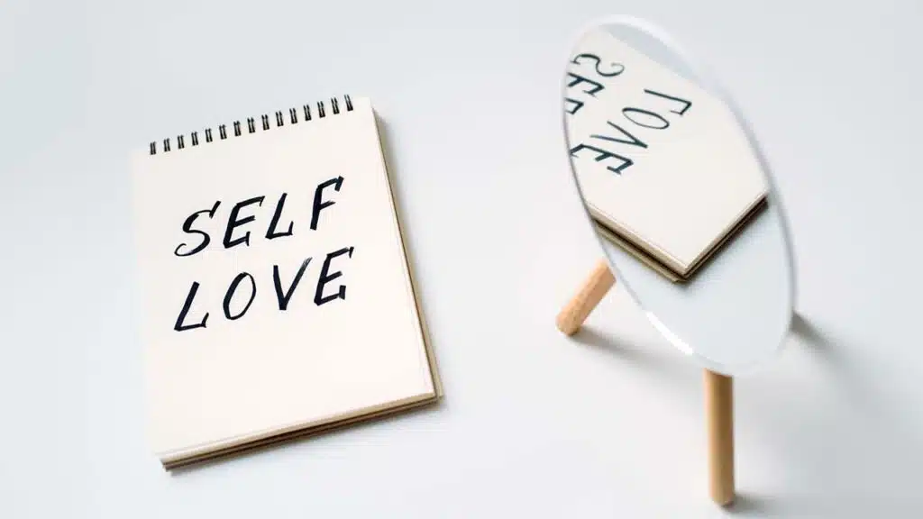 Notebook with 'Self Love' written on it, reflected in a small round mirror.