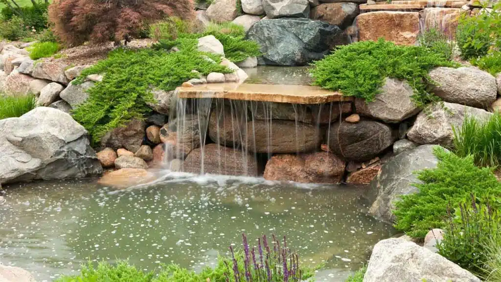A serene garden waterfall flows over layered stone slabs into a calm pond