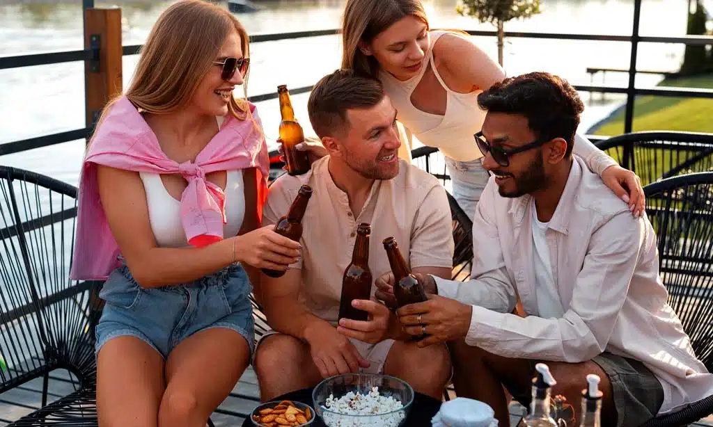 A group of four friends are sitting outdoors, enjoying a casual moment together, eating food and drinking alcohol by a waterfront.