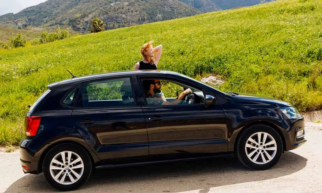 two people near a black hatchback car parked on a scenic road