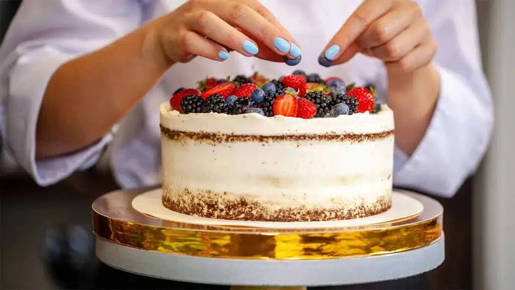 A person carefully decorating a cake with fresh berries, including strawberries, blackberries, and blueberries