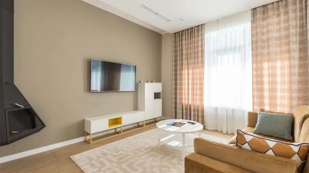 Modern living room with a wall-mounted TV, beige sofa, patterned curtains, and a round coffee table.