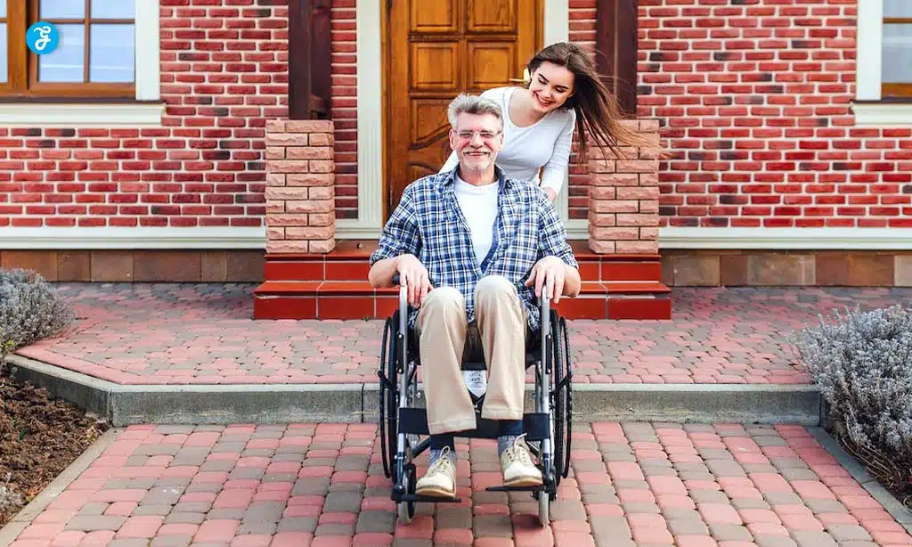 A smiling older man sits in a wheelchair, while a younger woman stands behind him, pushing the wheelchair