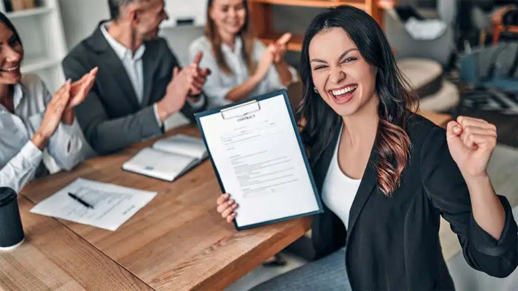Happy businesswoman celebrating while holding a signed contract as colleagues applaud during a meeting.
