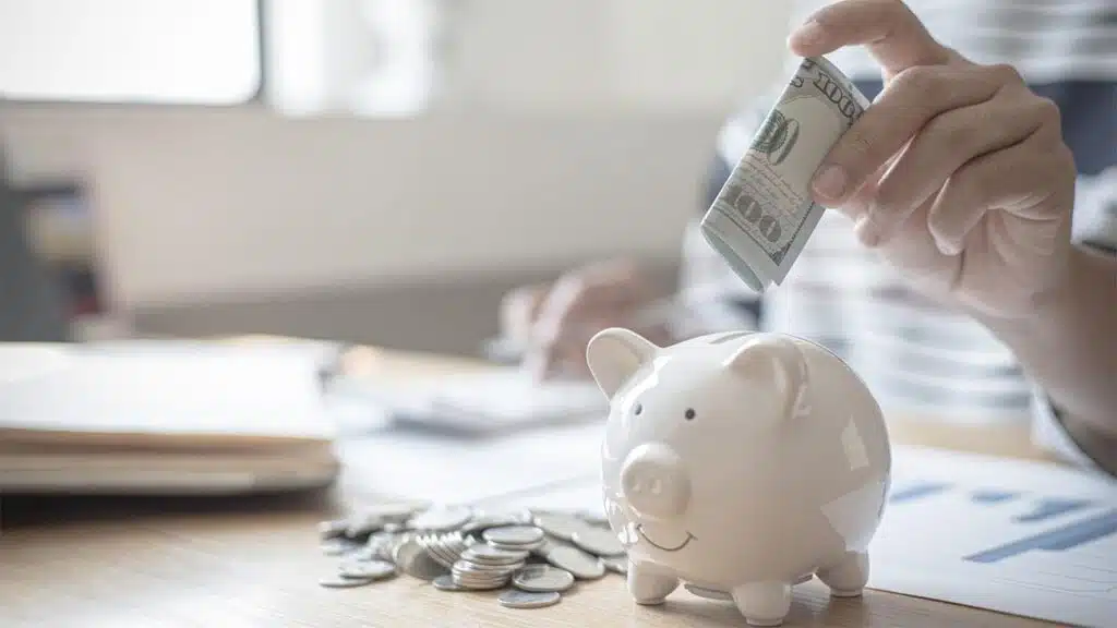 Person inserting a rolled-up US $100 bill into a white ceramic piggy bank