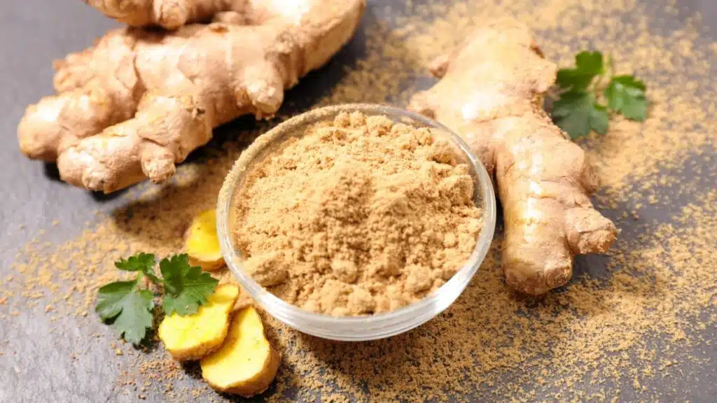 A small glass bowl filled with ground ginger powder, surrounded by fresh ginger roots