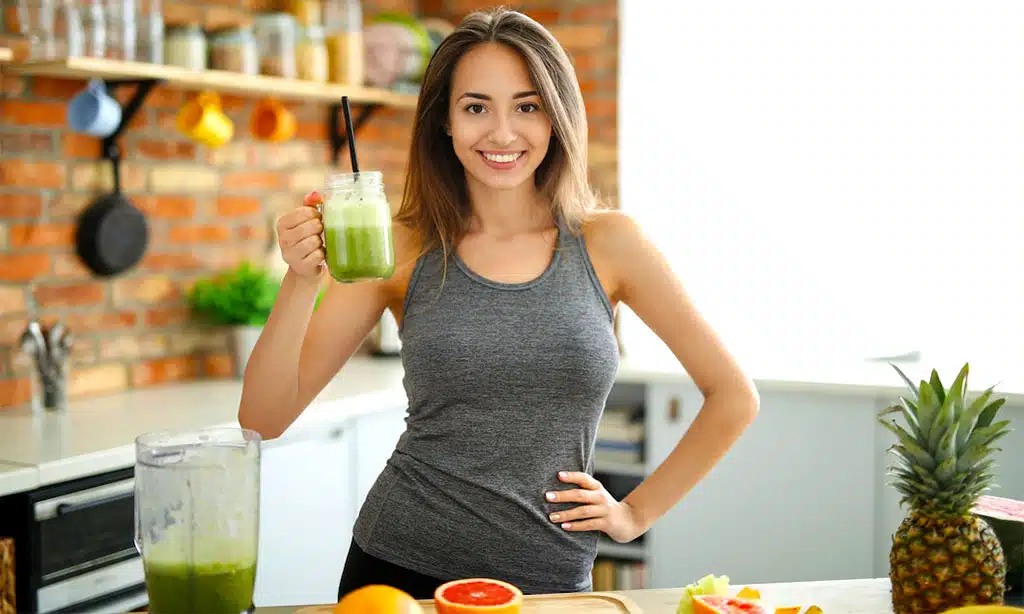 Smiling young woman in a modern kitchen holding a green smoothie, promoting a healthy lifestyle