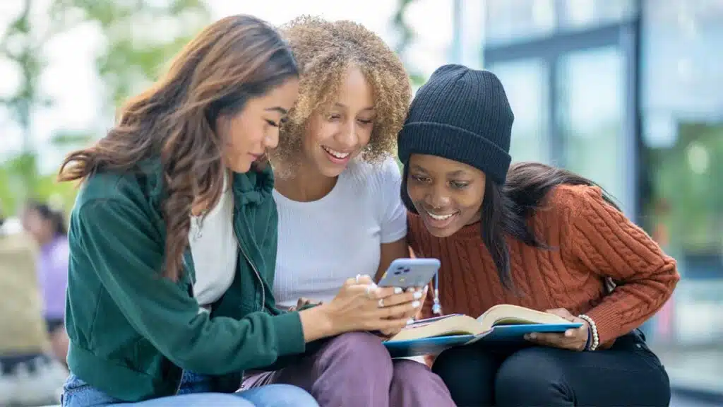 Three young women sitting on a bench, focused on a book they are reading together.