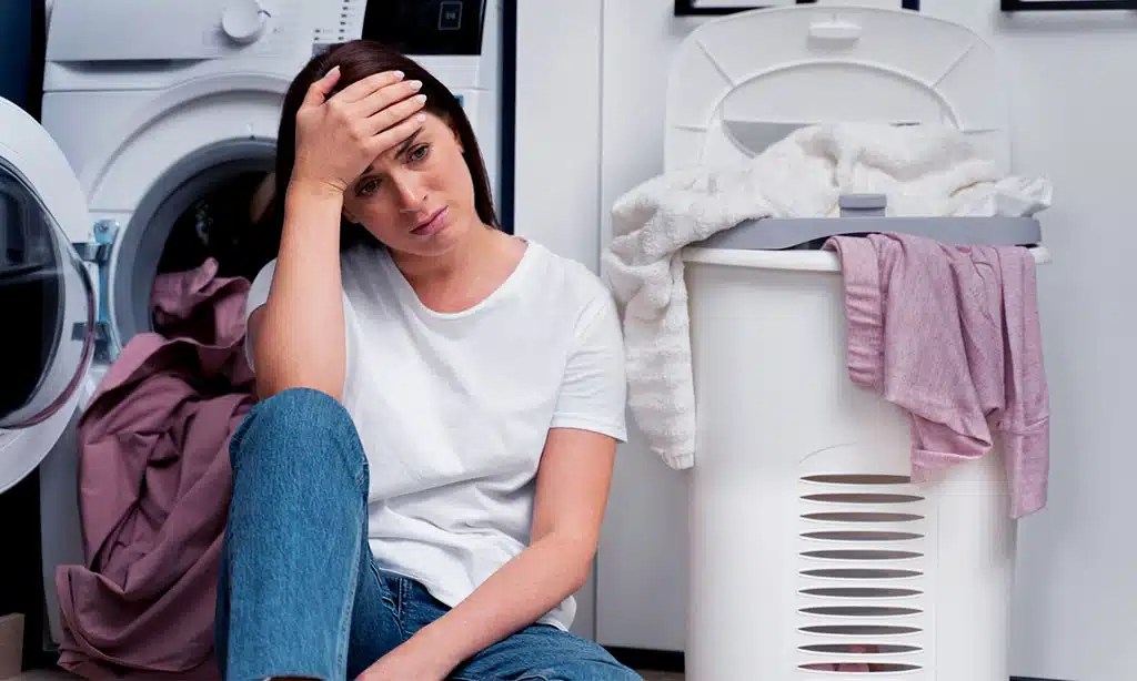 A woman sits on the floor, looking frustrated in front of a laundry machine with a full basket of clothes