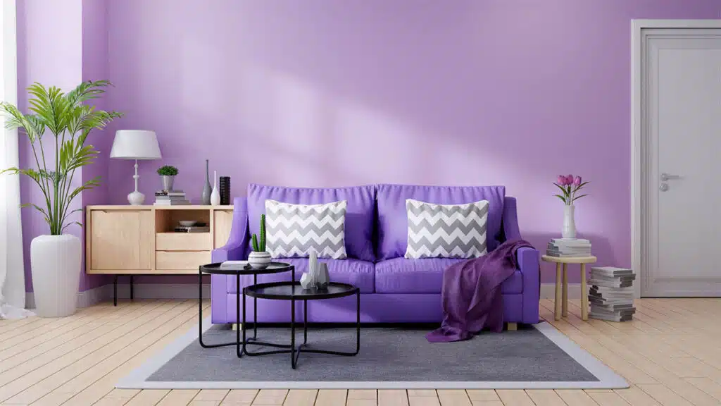 A lavender-colored wall complements a bold purple sofa, which is decorated with two gray and white chevron-patterned pillows