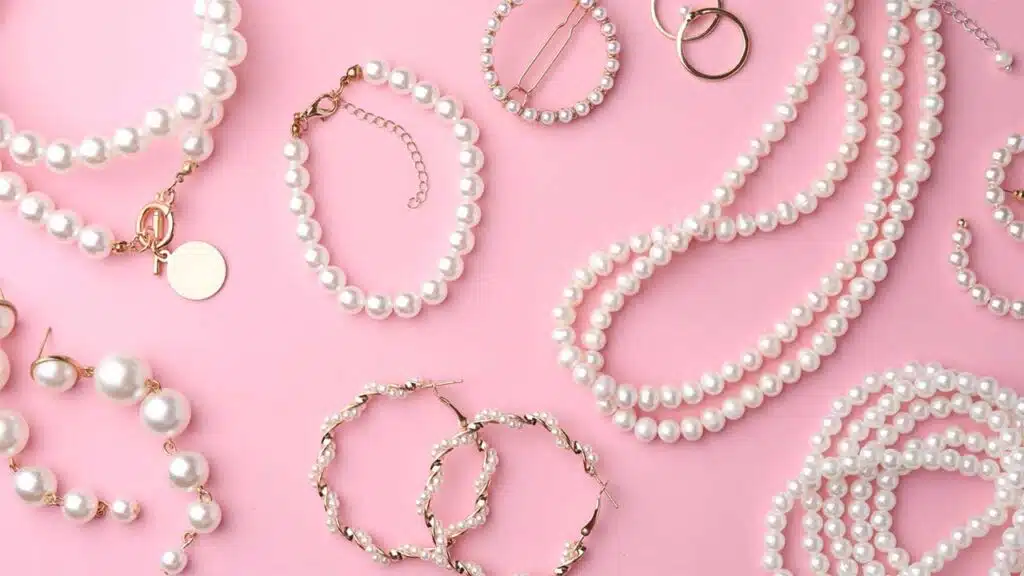 Elegant pearl jewelry set displayed on a pink background, featuring pearl necklaces, bracelets, earrings, and hair accessories with gold accents