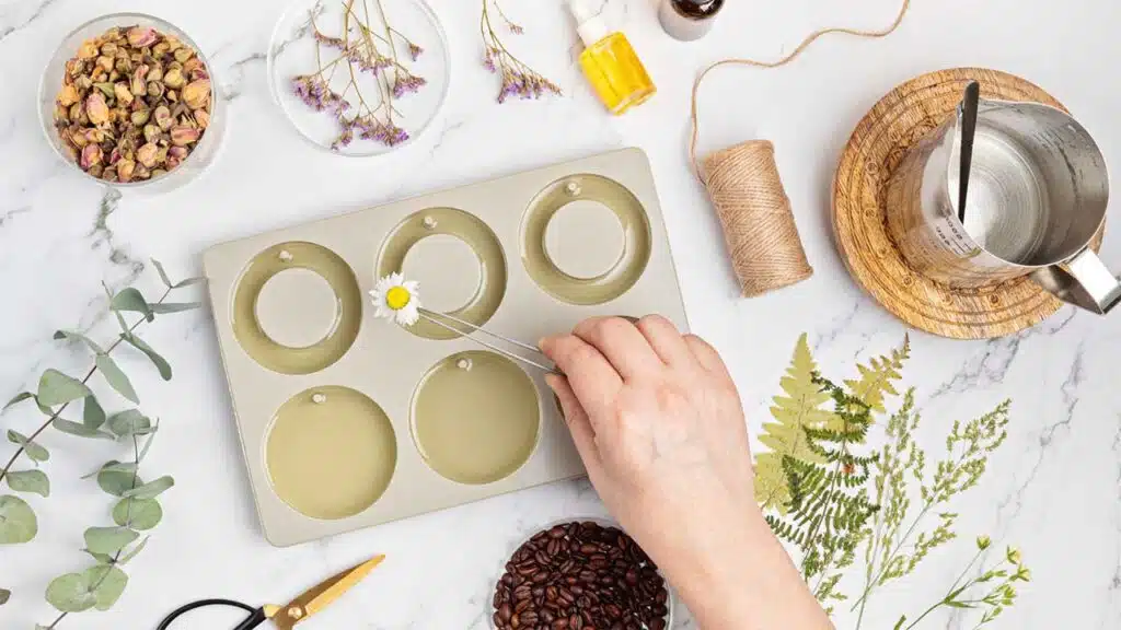 Person making homemade candles with dried flowers, essential oils, and natural ingredients on a crafting table.