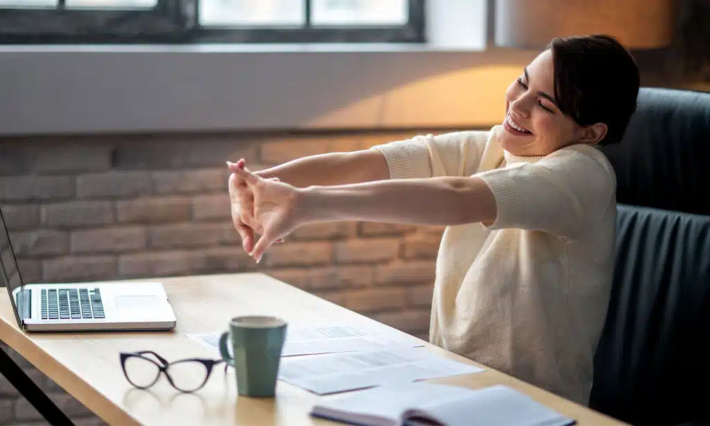 A woman sitting at her desk, taking a break from work. She is stretching her arms forward with a big smile on her face
