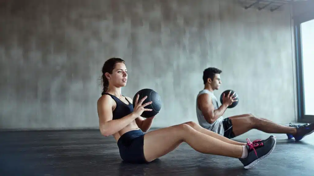 Two individuals working out together in a gym setting, performing an exercise using medicine balls.