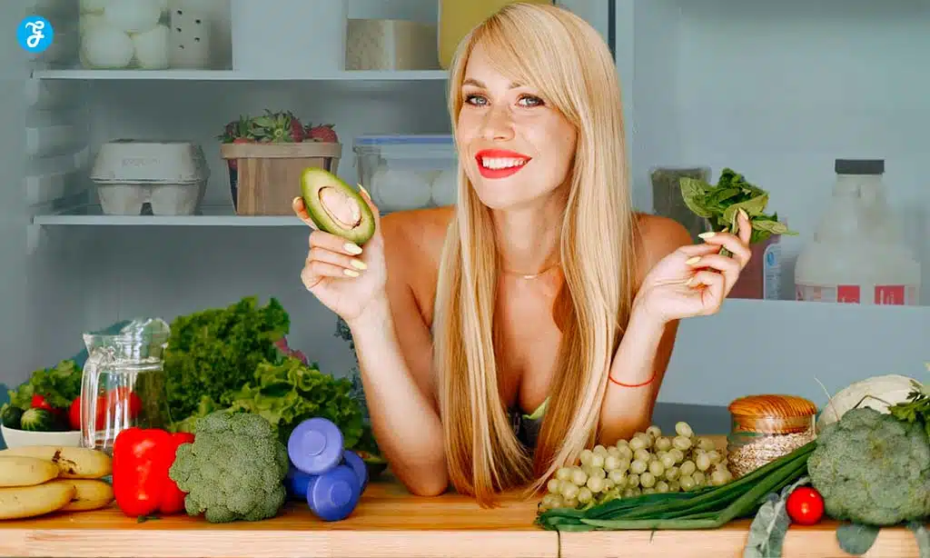 A smiling woman holds fresh avocado and spinach, surrounded by a colorful array of healthy foods.