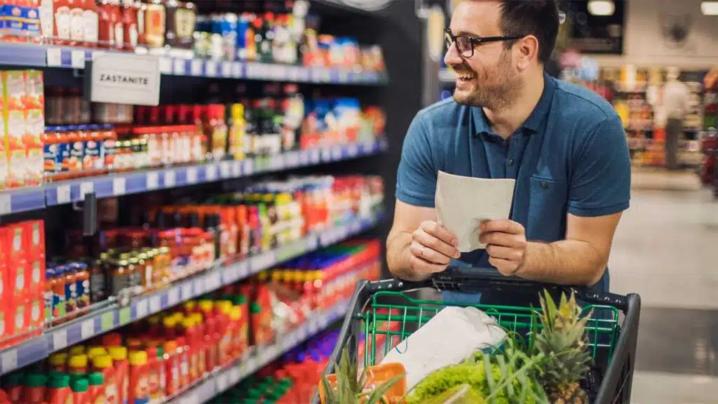 A man smiles while grocery shopping, pushing a cart filled with fresh produce and holding a shopping list.